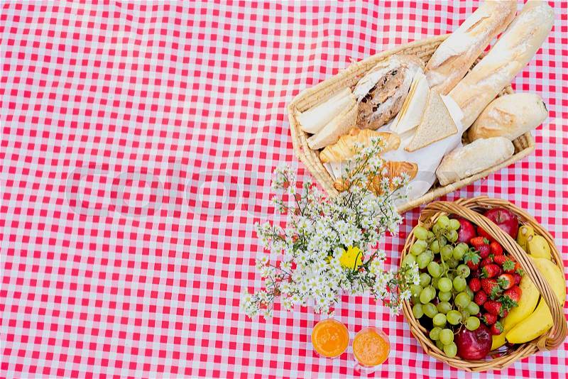 Picnic wicker basket with food, bread, fruit and orange juice on a red and white checked cloth in the field with green nature background. Picnic concept, stock photo