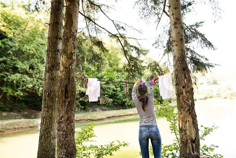 Beautiful woman on camping holiday in forest at the lake hanging clothes, rear view, stock photo