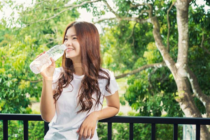 Happy smiling asian woman holding water bottle to drink, relaxing on her balcony with blurred trees in background and space for text or copy space, good for healthy lifestyle theme concept, stock photo