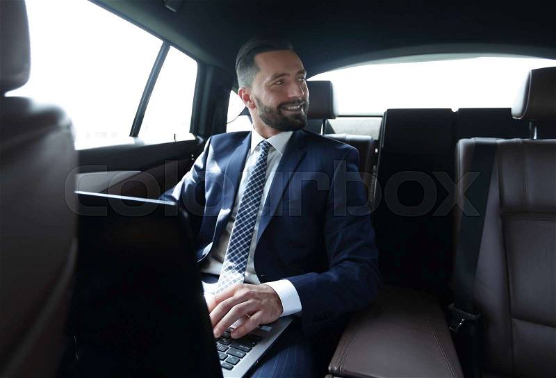 Successful businessman with laptop sitting in the back seat in the car, stock photo