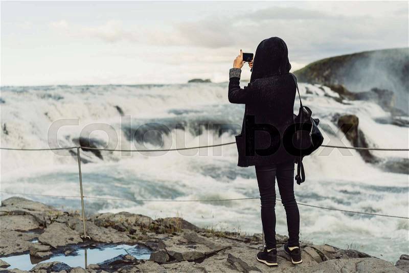 Back view of young woman photographing majestic waterfall in Iceland , stock photo