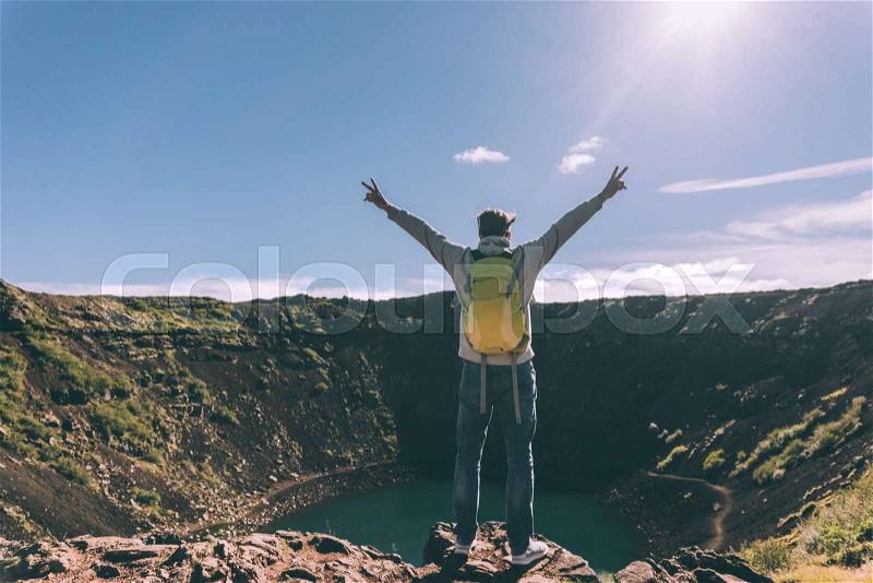 Back view of young man with backpack standing with raised hands and looking at majestic crater lake in Iceland, stock photo