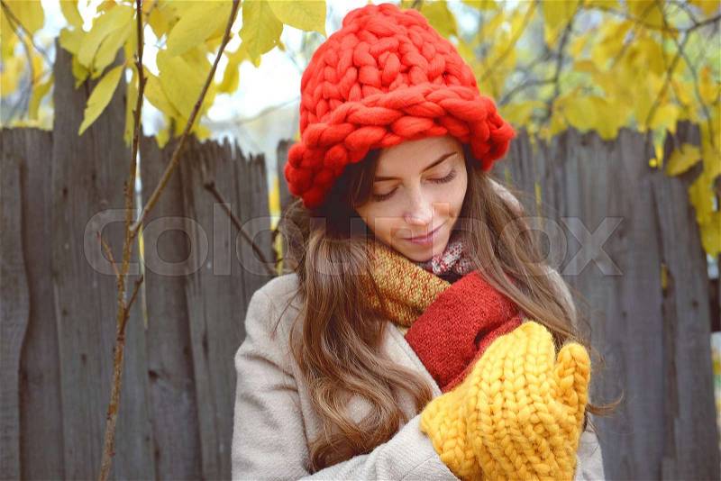 Colorful fall scenery. Young woman in beige wool coat and large knitted red hat and yellow mittens walking outdoorin countryside, stock photo