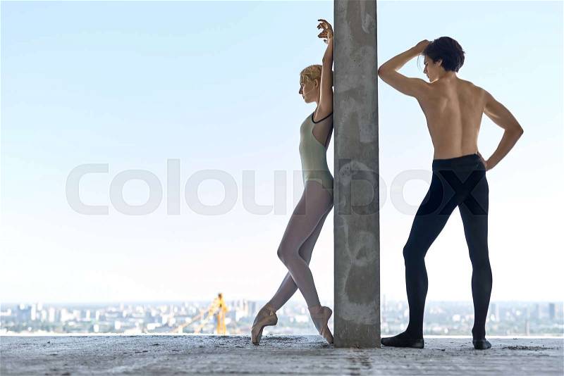 Athletic couple of ballet dancers lean on the concrete wall on the floor of the unfinished building on the cityscape background. Topless guy wears a black dance pants, girl wears a green leotard, stock photo