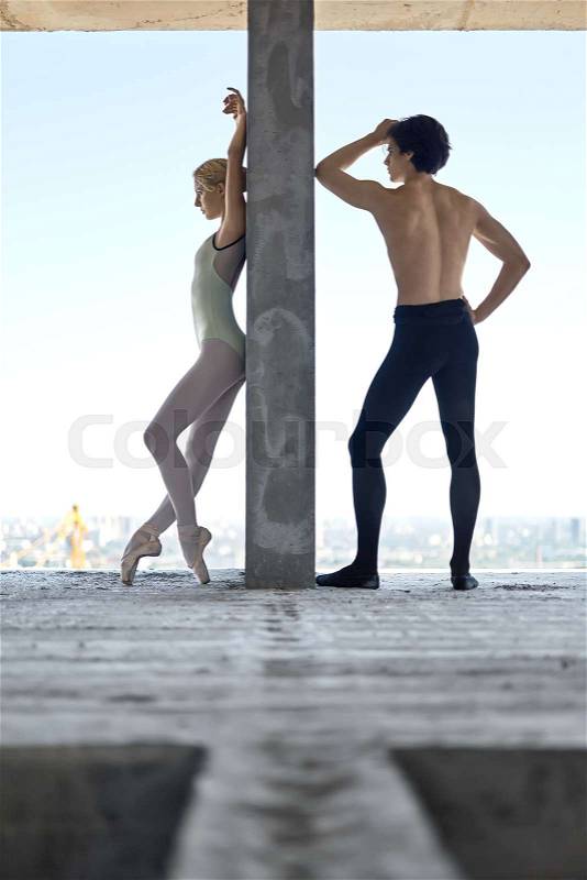 Attractive couple of ballet dancers lean on the concrete wall on the floor of the unfinished building on the cityscape background. Topless guy wears a black dance pants, girl wears a green leotard, stock photo