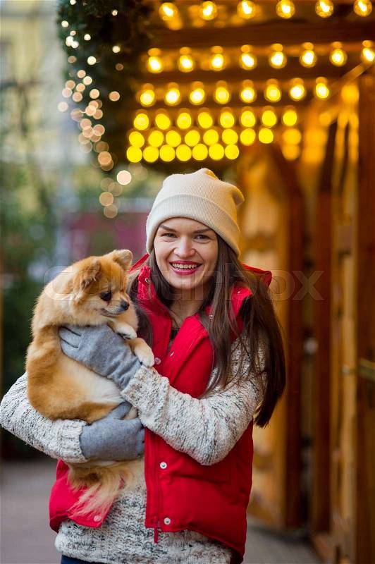 Cheerful young woman with a pet at the Christmas bazaar. Owner holds of a little pomeranian spitz. Woman has a festive mood, stock photo