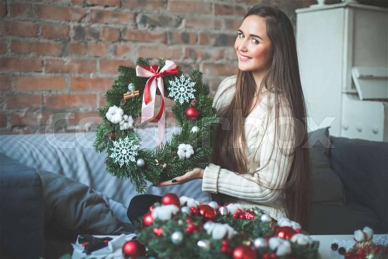 Young Cute Woman with Christmas Evergreen Tree Wreath with Red Glass balls, Cones, Ribbon and Cinnamon at Home, stock photo