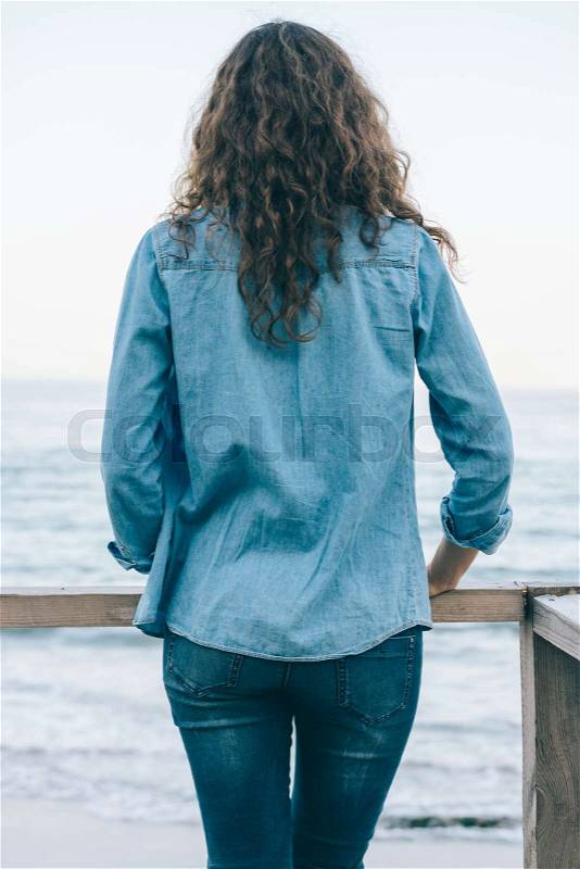 Slender woman with curly hair in denim clothes looks at the sea on the beach, stock photo