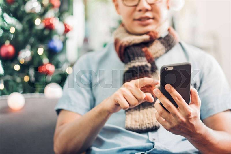 Asia man wear santa hat use mobile call friends and sitting on sofa at Christmas tree in house,invite people to come winter festive holiday party, stock photo