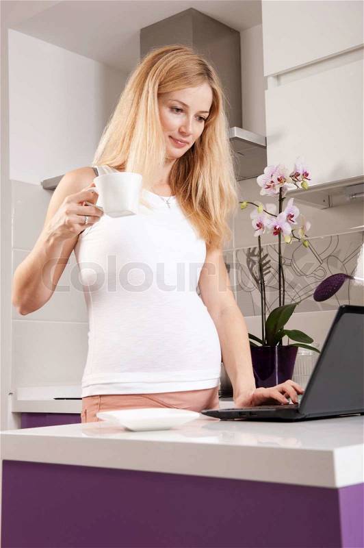 Young pregnant blonde woman drinking tea in kitchen and surfing the Internet, stock photo