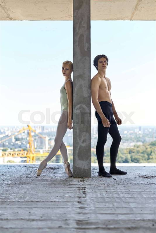 Serious couple of ballet dancers lean on the concrete wall on the floor of the unfinished building on the cityscape background. Topless guy wears a black dance pants, girl wears a green leotard, stock photo