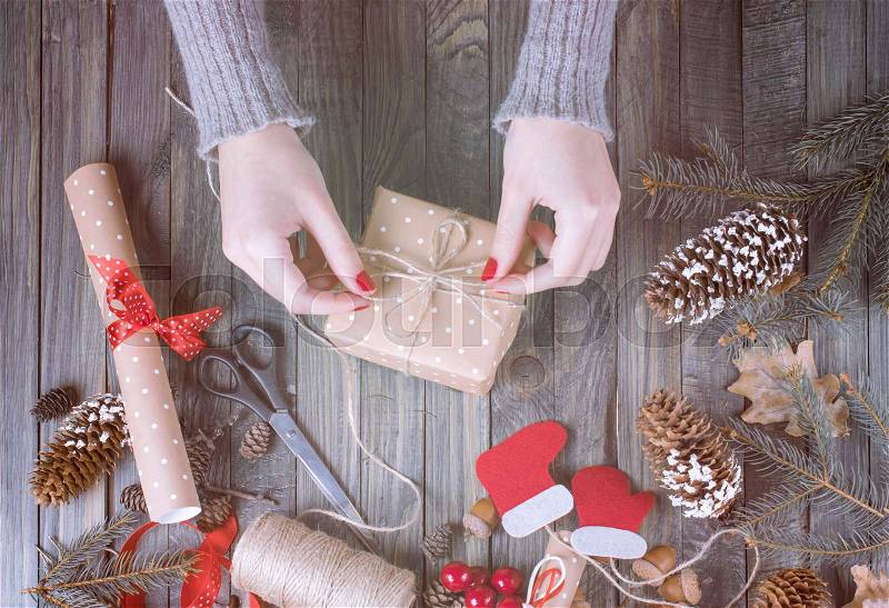 Christmas background. Top view of woman hands wrap New Year present and tie bow. Packed gifts and scrolls, spruce branches and tools on shabby wooden table, stock photo