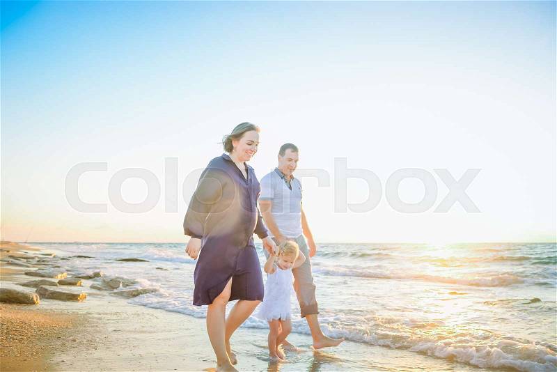 Happy family of three - pregnant wife, father and daughter having fun walking on beach at sunset. Family traveling concept. Backlight, soft selective focus. Copy space, stock photo
