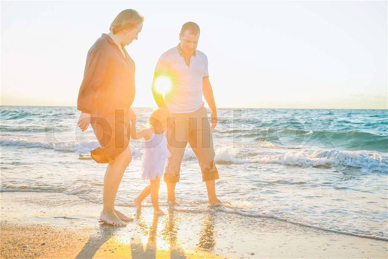 Happy family of three - pregnant wife, father and daughter having fun walking on beach at sunset. Family traveling concept. Backlight, soft selective focus. Copy space, stock photo
