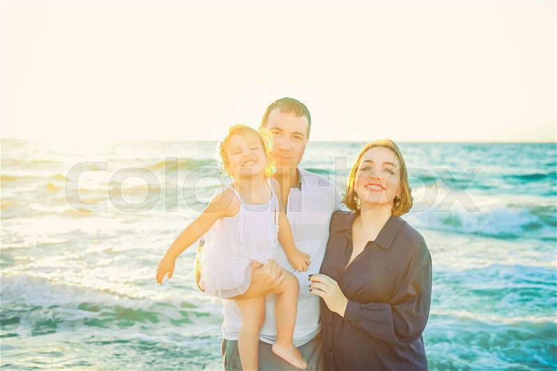 Portraits of happy family of three - pregnant wife, father and daughter having fun walking on beach at sunset. Family traveling concept. Backlight, soft selective focus. Copy space, stock photo