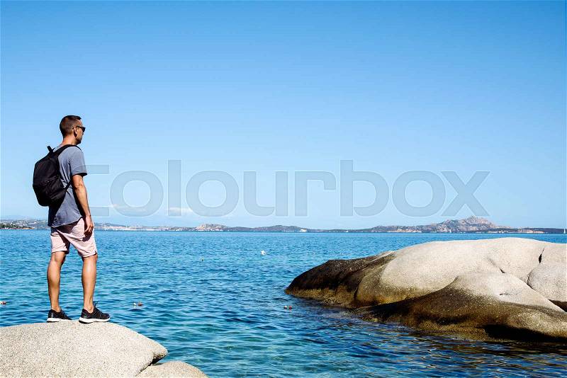 A young caucasian man, seen from behind, carrying a backpack observing the sea in the Costa Smeralda, in Sardinia, Italy, with a large blank space in the sky, stock photo