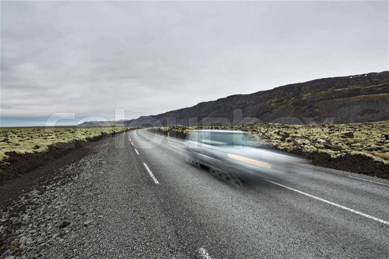 Roadway with orange roadside pillars between the green fields and mountains on the background of the cloudy sky in Iceland. There is a blurry car in motion on the driveway. Horizontal, stock photo
