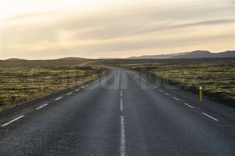 Textured roadway with orange roadside pillars between the green fields on the background of the mountains and cloudy sunset sky in Iceland. Horizontal, stock photo
