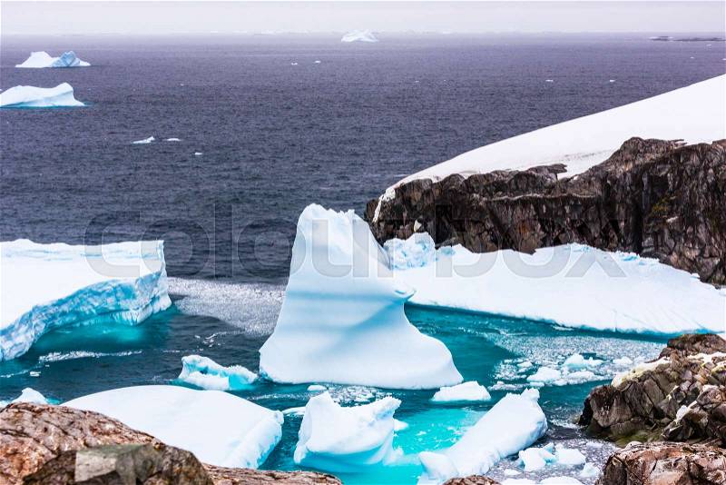 Cold still waters of antarctic sea lagoon with drifting blue icebergs among rocky cliffs of Peterman island, Antarctica, stock photo