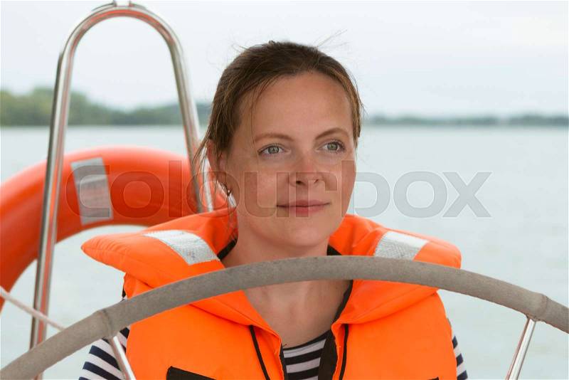 Portrait of happy young woman in life jacket with steering wheel on the yacht, stock photo