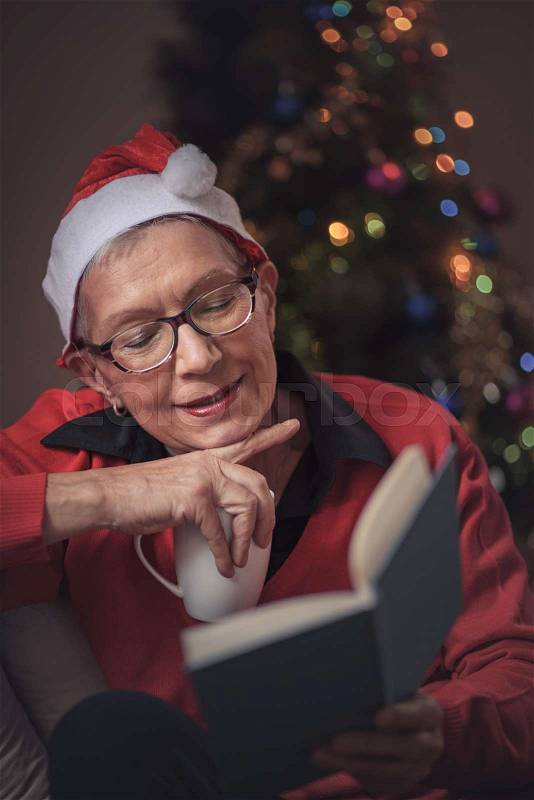 Senior old lady reading her favorite book during December holiday, house decorated with a christmas tree, she's wearing a christmas hat, stock photo