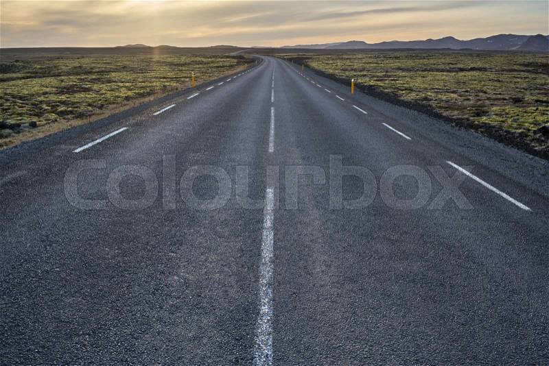 Route with orange roadside pillars between the green fields on the background of the mountains and cloudy sunset sky in Iceland. Horizontal, stock photo