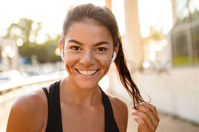Close up portrait of a smiling fitness woman in earphones looking at camera while standing outdoors, stock photo