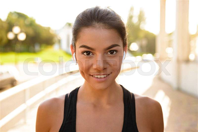 Close up portrait of a young fitness woman in earphones looking at camera while standing outdoors, stock photo