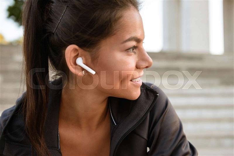 Close up portrait of a smiling fitness woman in earphones looking away while sitting outdoors, stock photo