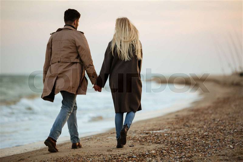 Back view of a young couple in love holding hands while walking along the beach, stock photo