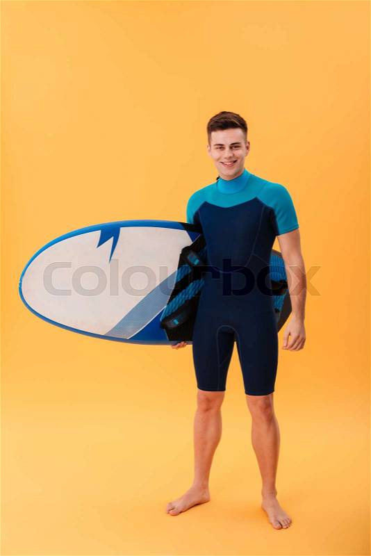 Full length photo of young smiling man in swimsuit holding surfing board while standing and looking at camera, isolated on yellow background, stock photo
