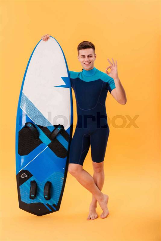 Full length photo of young attractive man in swimsuit holding surfboard while standing and showing OK gesture, isolated over yellow background, stock photo