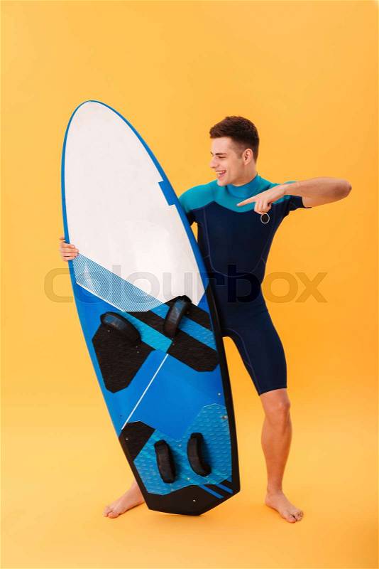 Full length photo of cheerful young man in swimsuit pointing with finger on his surfboard, isolated on yellow background, stock photo