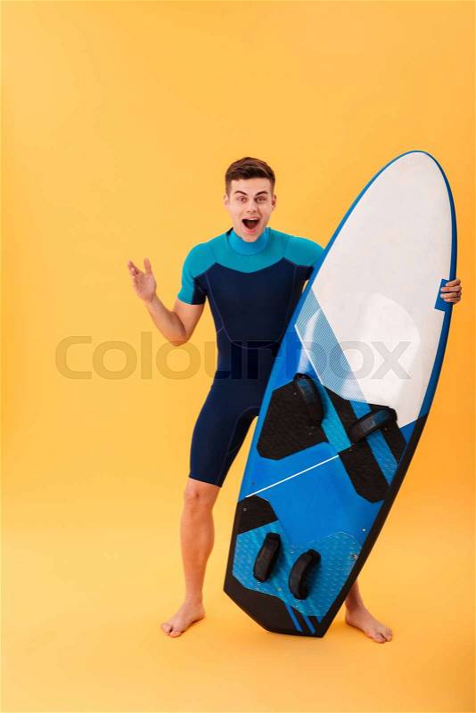 Full length photo of happy exited brunette man in swimsuit holding surfbourd while standing and lookin at camera, isolated over yellow background, stock photo