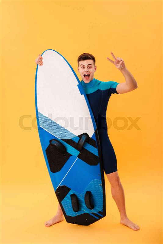 Full length photo of happy young man in swimsuit showing peace gesture while standing and holding surfboard, isolated on yellow background, stock photo