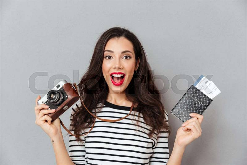 Portrait of an excited woman holding vintage camera and passport with flying tickets isolated over gray background, stock photo