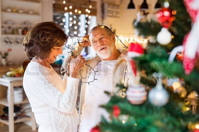 Beautiful senior couple in white woolen sweaters decorating Christmas tree inside their house. Man and woman having fun, stock photo