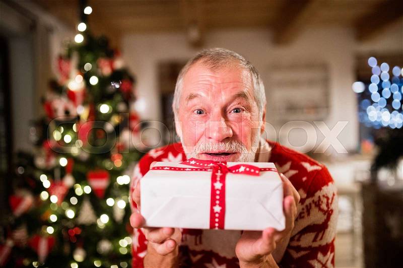 Senior man standing in front of illuminated Christmas tree inside his house holding a present, stock photo