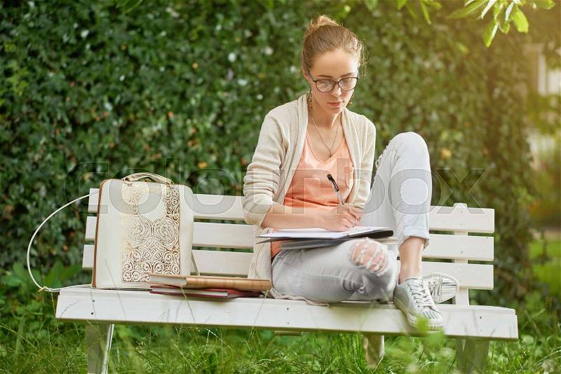 Young girl looks like a student, writes something on the white park s bench. There are some books and a stylish backpack near her. She wears an eyeglasses, blue jeans, light t-shirt and a cardigan, stock photo