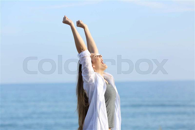 Side view portrait of an excited woman screaming and raising arms on the beach with the sea in the background, stock photo