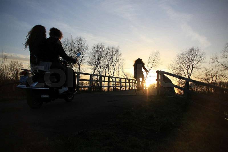 Two schoolgirls on the moped and one schoolgirl is biking and going home over the wooden bridge at the countryside at sunset in the soft winter, stock photo