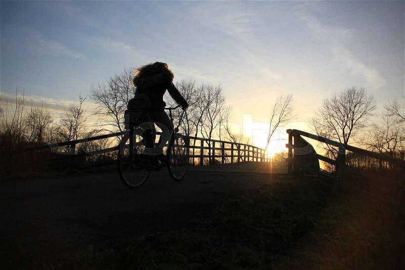 The solitary schoolgirl is biking over the wooden bridge and is going home at the countryside at sunset in the soft winter, stock photo