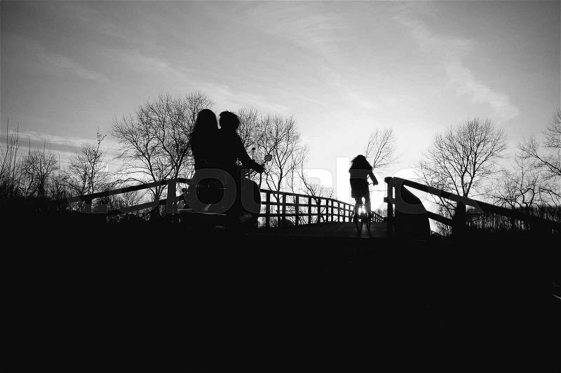 Two schoolgirls on the moped and one schoolgirl is biking and going home over the wooden bridge at the countryside at sunset in the soft winter in black and white, stock photo