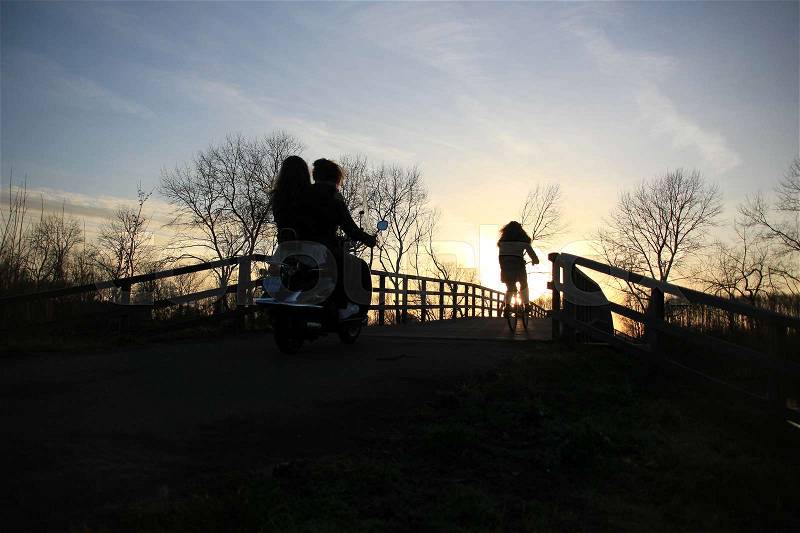 Two schoolgirls on the moped and one schoolgirl is biking and going home over the wooden bridge at the countryside at sunset in the soft winter, stock photo