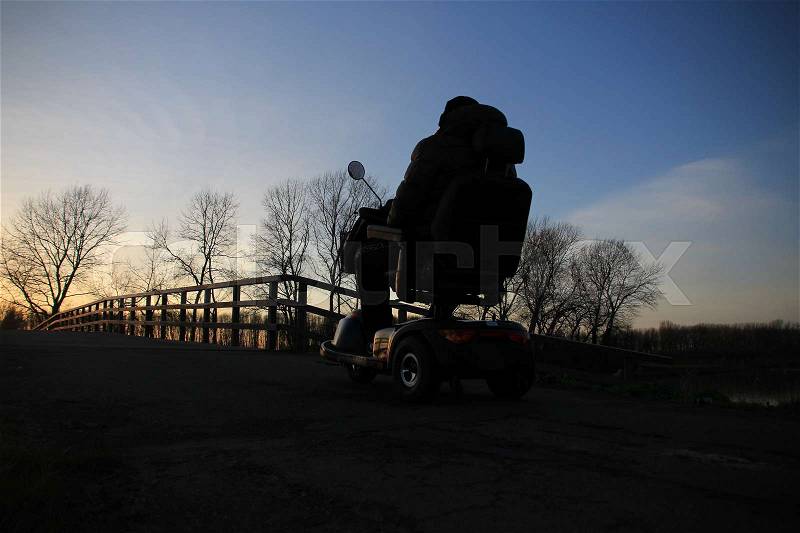 The man in the mobility scooter is going home over the wooden bridge at the countryside at sunset in the soft winter, stock photo
