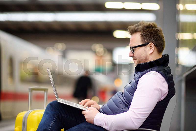 Freelancer working with a laptop in a train station while is waiting for transport. Handsome middle age man on on railway station platform, stock photo