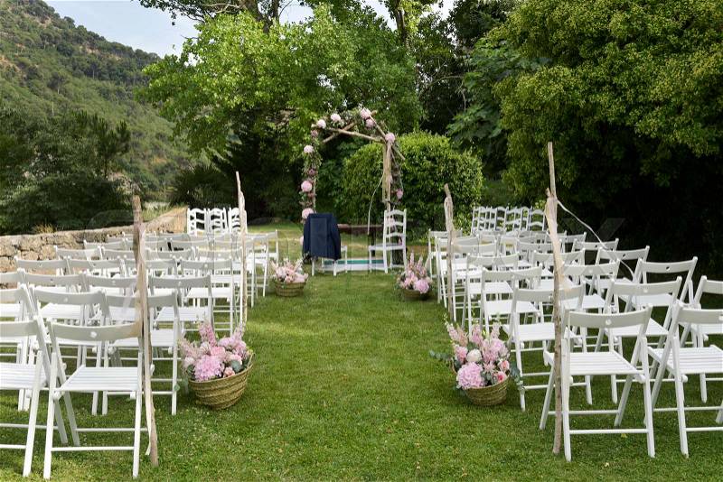 A view of the scene of an open-air wedding ceremony, with some rows of white folding chairs and some arrangements of different pale pink flowers, and a floral arch in the background, stock photo