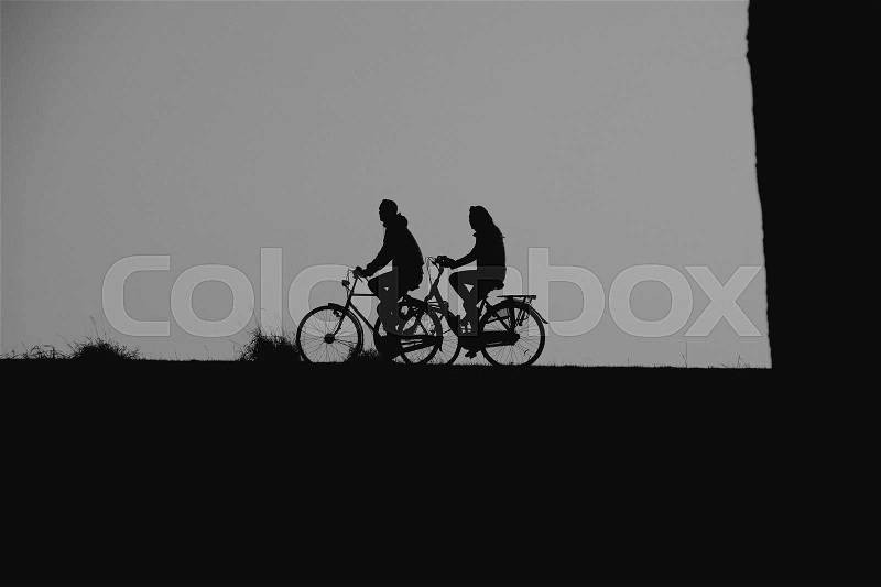A big trunk and a couple, man and wife are biking on the dyke in the village Hellevoetsluis at sunset in the summer in black and white, stock photo