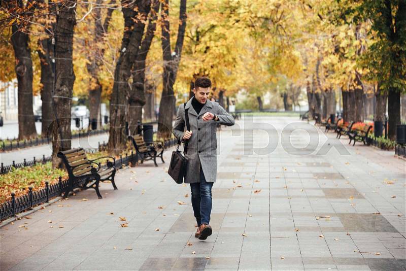 Picture of good-looking caucasian man in coat with bag strolling in city park, and looking at his watch, stock photo