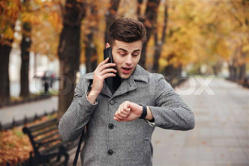 Picture of young guy talking on smartmobile while looking on his watch, being late, stock photo